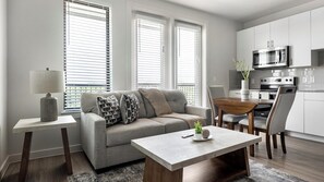 Ein modernes Wohnzimmer mit Sofa, Couchtisch und Esstisch. Große Fenster mit Jalousien, eine Lampe und eine Küchenzeile mit Geräten.
