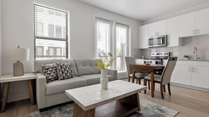 A modern living room with a sofa, a coffee table, and a dining area with chairs. There is a kitchen with white cabinets and appliances. - Landing | Modern 1BD, Gym, Pool (Austin)
