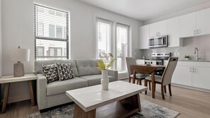A modern living room with a sofa, a coffee table, and a dining area with chairs. There is a kitchen with white cabinets and appliances. - Landing | Stunning 1BD, Pool, Gym (Austin)