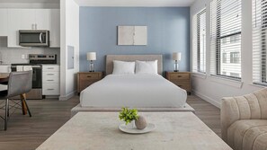 Une chambre moderne avec un grand lit, des tables de chevet, une table d’appoint, une table basse avec une plante et une vue sur la cuisine.