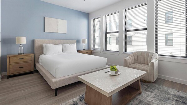 Une chambre à coucher moderne, avec un lit, des tables de chevet, une chaise, une table basse et de grandes fenêtres équipées de stores.