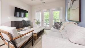 Un salon comprenant un canapé, une table basse, un téléviseur et un balcon aménagé avec du mobilier d’extérieur.