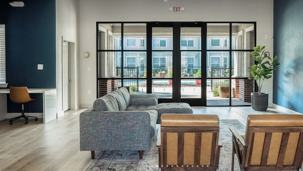 Un intérieur moderne avec un canapé d’angle, deux fauteuils en cuir, une plante en pot et une vue sur l’extérieur, accessible par de grandes portes vitrées.