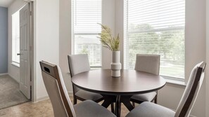 A dining area with a round table, four chairs, a vase with greenery, and a window with blinds.