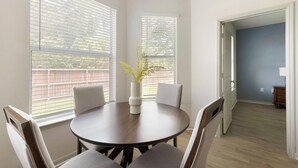 Een eetgedeelte met een ronde tafel, vier stoelen, een vaas met bloemen en grote ramen met jaloezieën.