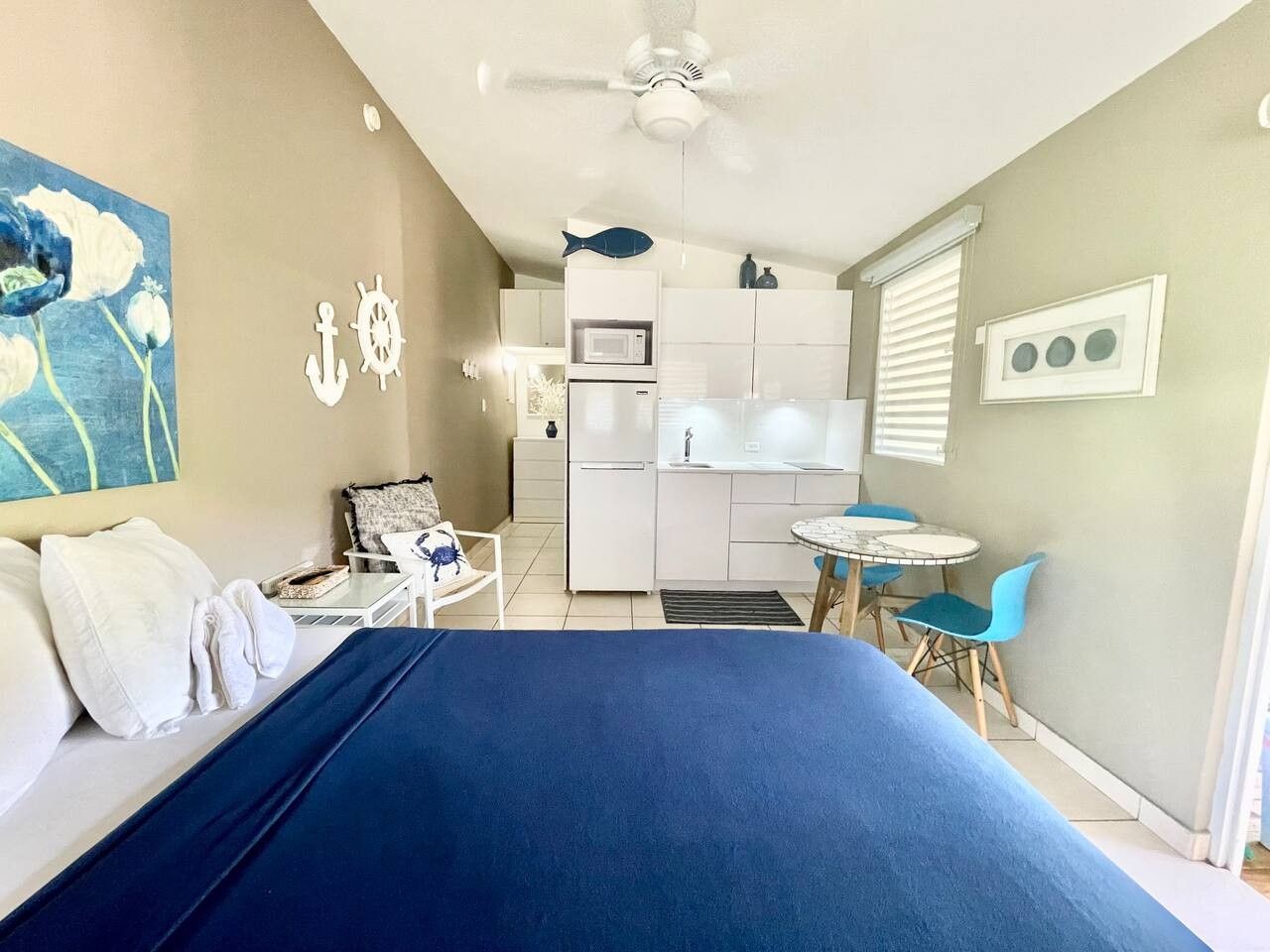 Une chambre d’hôtel compacte comprenant un lit, une petite kitchenette, un coin repas avec une table et des chaises, et un ventilateur de plafond.