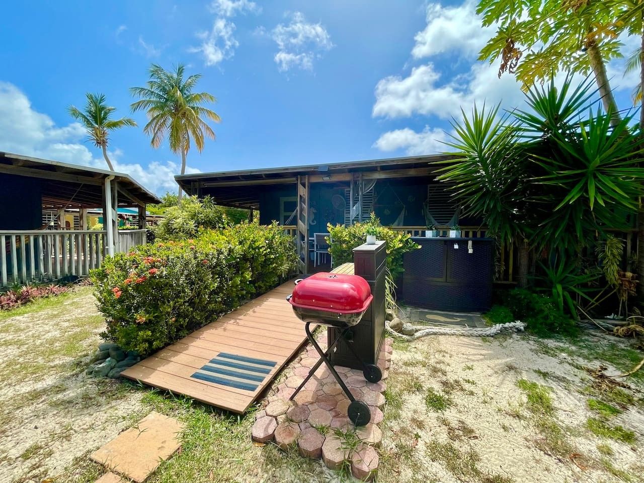 Un espace extérieur aménagé avec un barbecue, une terrasse en bois et un petit jardin agrémenté de palmiers et d’arbustes.
