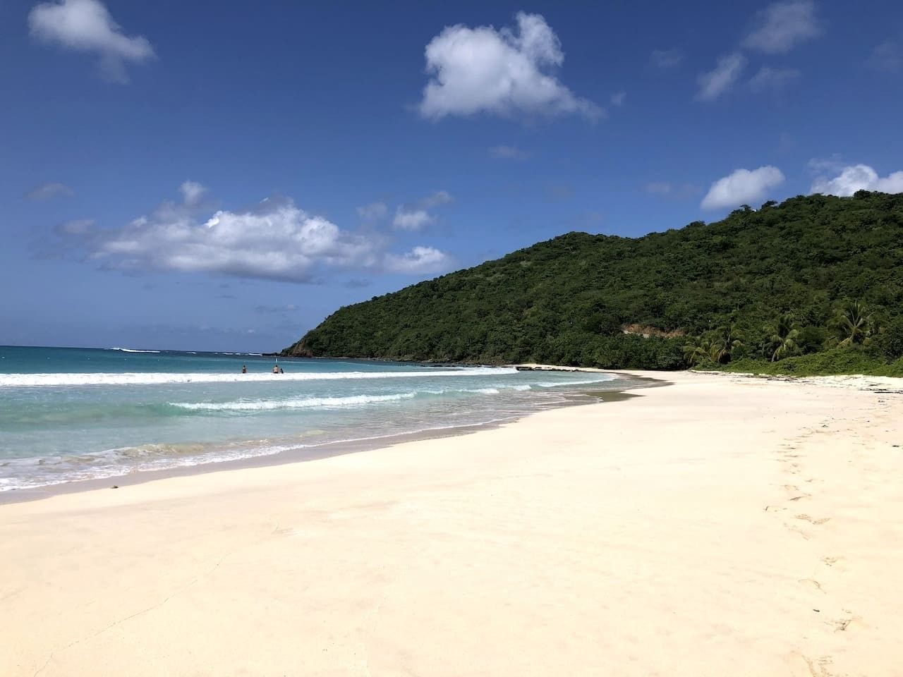 Una playa con aguas azul turquesa, una orilla arenosa y una colina boscosa al fondo.