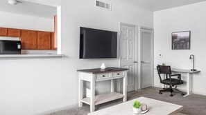 A modern living room with a flat-screen TV, a desk with a chair, a coffee table, and a kitchen area with wooden cabinets and a black refrigerator. - Landing | Incredible 2BD, Yoga Studio, Pool (Humble)