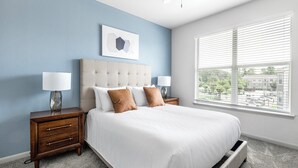A bedroom with a bed, two bedside tables with lamps, a headboard, a window with blinds, and a wall-mounted artwork.