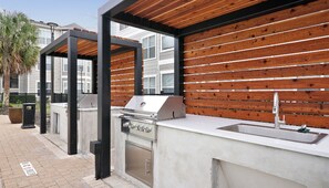 An outdoor kitchen area with a grill, sink, and wooden panel backdrop. - Landing | Beautiful 2BD, Pool, Yoga Studio (Humble)