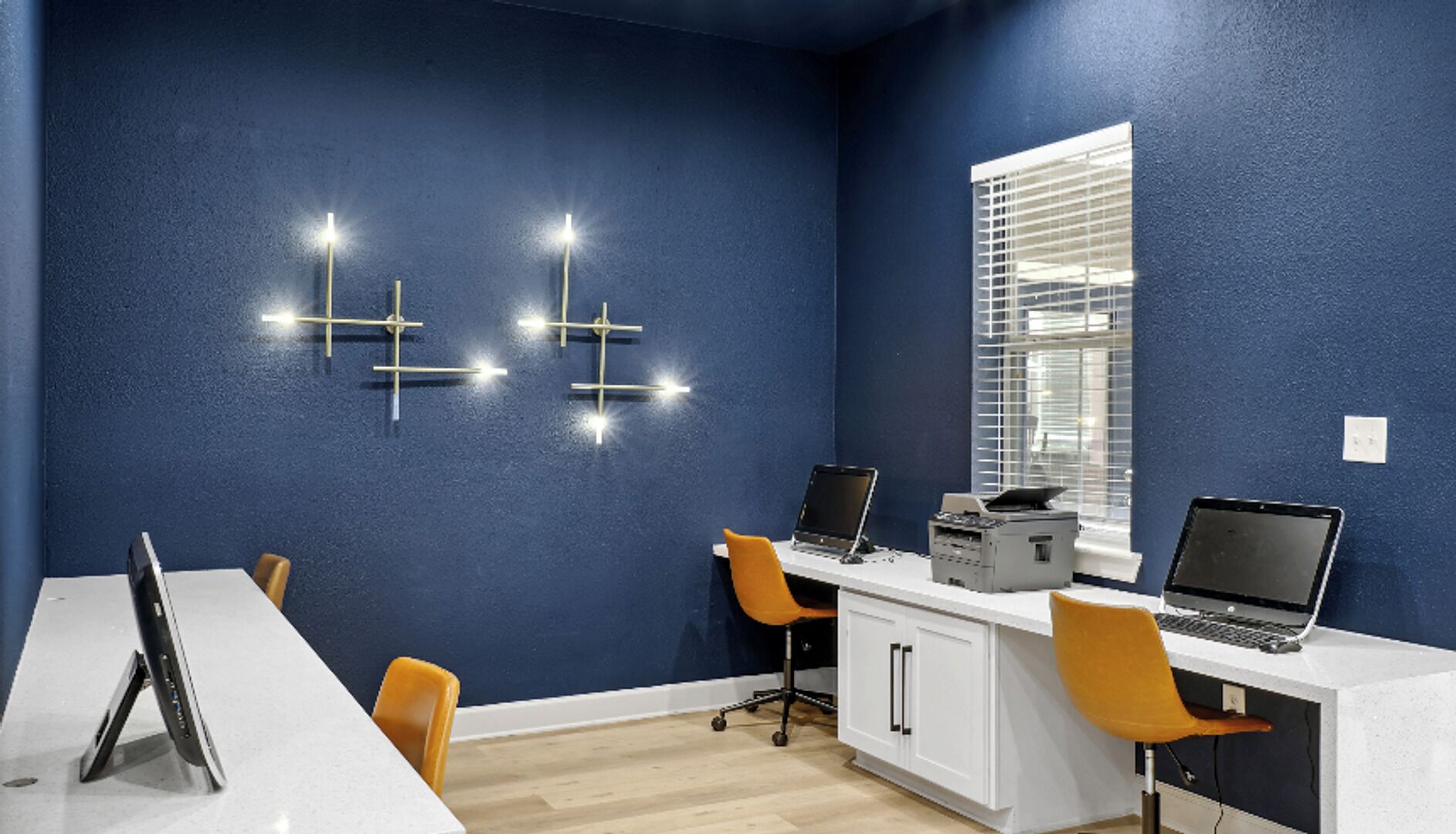A conference room with a white table, two chairs, a desk with two laptops, a printer, and a wall-mounted light fixture.