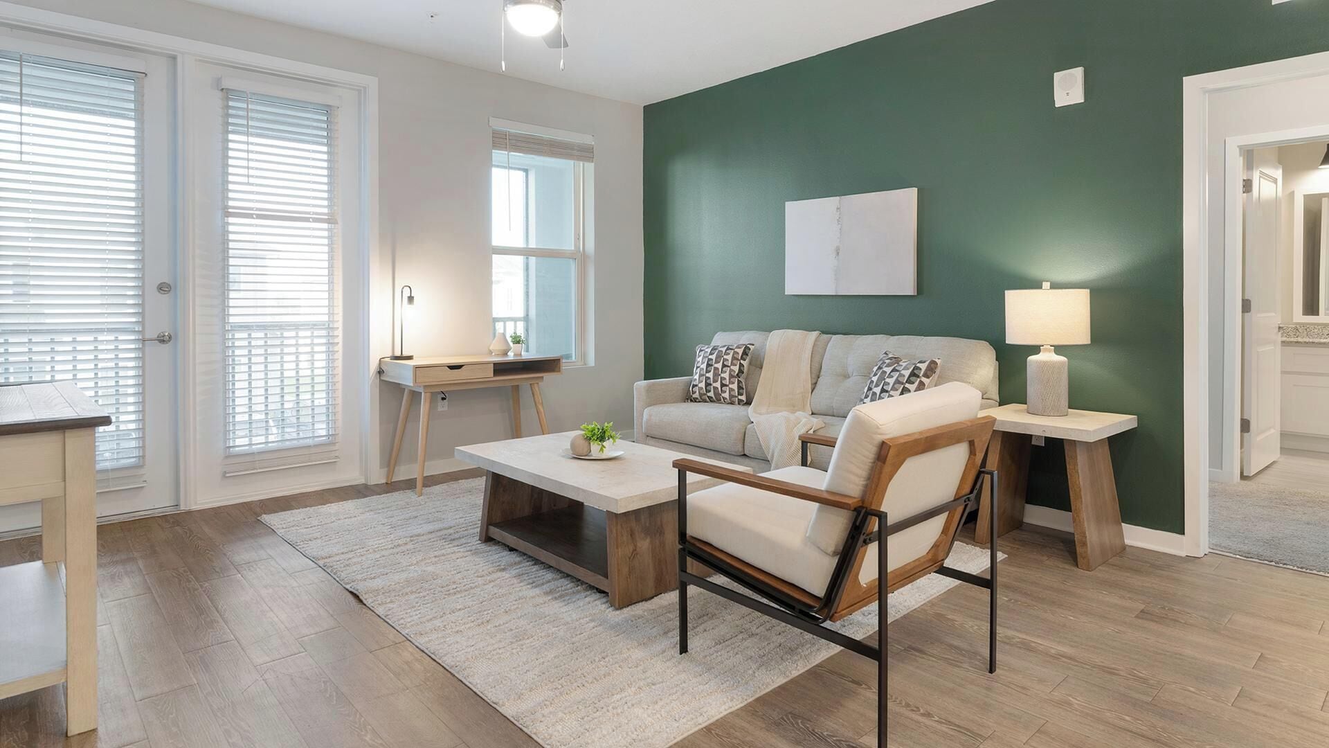 A living room with a sofa, coffee table, and lamp, a desk with a chair, and a green accent wall.