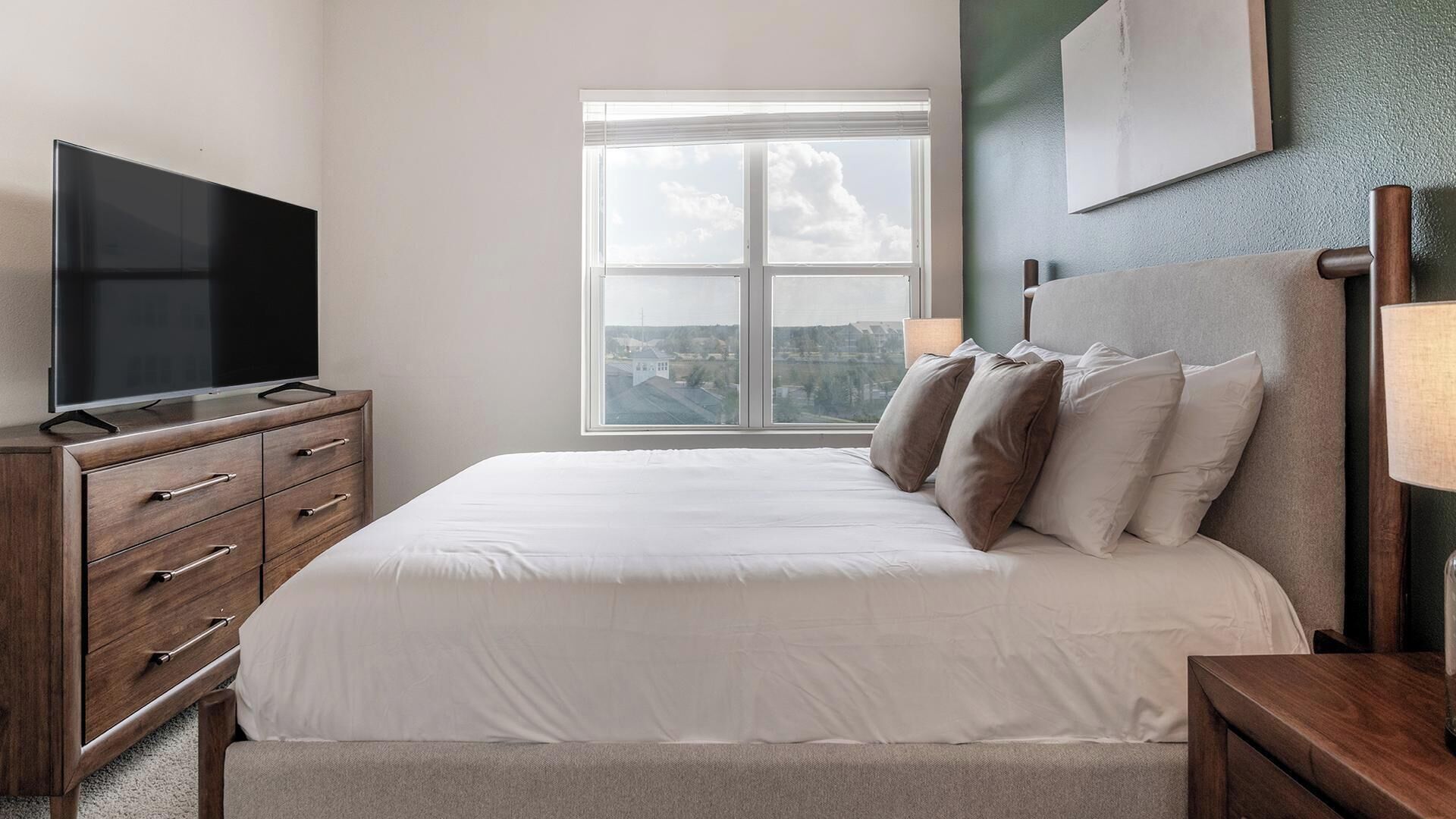 A bedroom with a bed, a TV on a dresser, a window with a view, and a lamp on a nightstand.