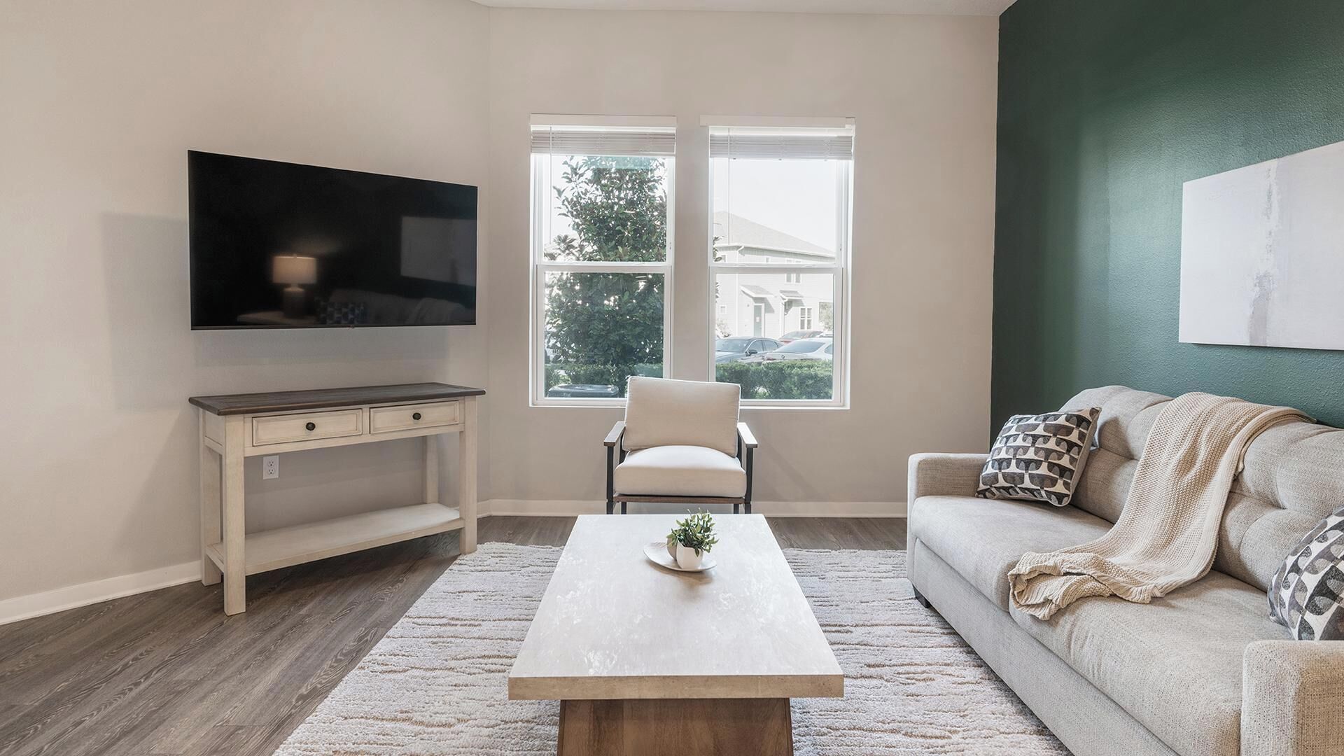 Una sala de estar con televisor de pantalla plana, un sofá blanco, una mesa de centro, una silla y una ventana con vistas a una casa y árboles.