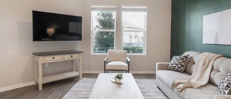 Una sala de estar con televisor de pantalla plana, un sofá blanco, una mesa de centro, una silla y una ventana con vistas a una casa y árboles.