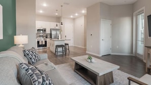 A modern living room with a sofa, coffee table, and kitchen in the background. - Landing | Stylish 1BD, Yoga Studio, Gym (Four Corners)