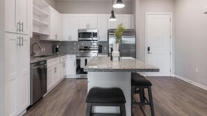 A modern kitchen with white cabinets, stainless steel appliances, a granite countertop, and a central island with stools. - Landing | Stylish 1BD, Yoga Studio, Gym (Four Corners)