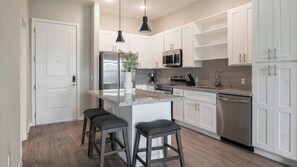 A modern kitchen with white cabinetry, stainless steel appliances, a granite countertop, and two bar stools. - Landing | Incredible 1BD, Gym, Yoga Studio (Four Corners)