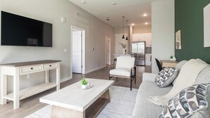 A modern living room with a flat-screen TV, a white sofa, a coffee table, and a dining area with a kitchen in the background. - Landing | Spectacular 1BD, Yoga Studio, Gym (Four Corners)