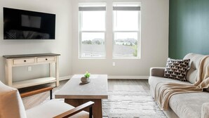 A living room with a sofa, a coffee table, a TV mounted on the wall, and a window with a view of buildings. - Landing | Spectacular 1BD, Yoga Studio, Gym (Four Corners)