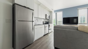 Eine moderne Küche mit Kühlschrank, Mikrowelle und Backofen, ein graues Sofa und ein Fernseher in einem Wohnbereich mit Fenster.