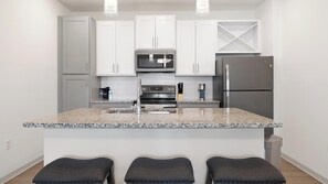 A modern kitchen with white cabinets, a stainless steel refrigerator, a microwave, and a granite countertop. There are three bar stools at the island. - Landing | Spectacular 1BD, Game Room (Lake Wales)