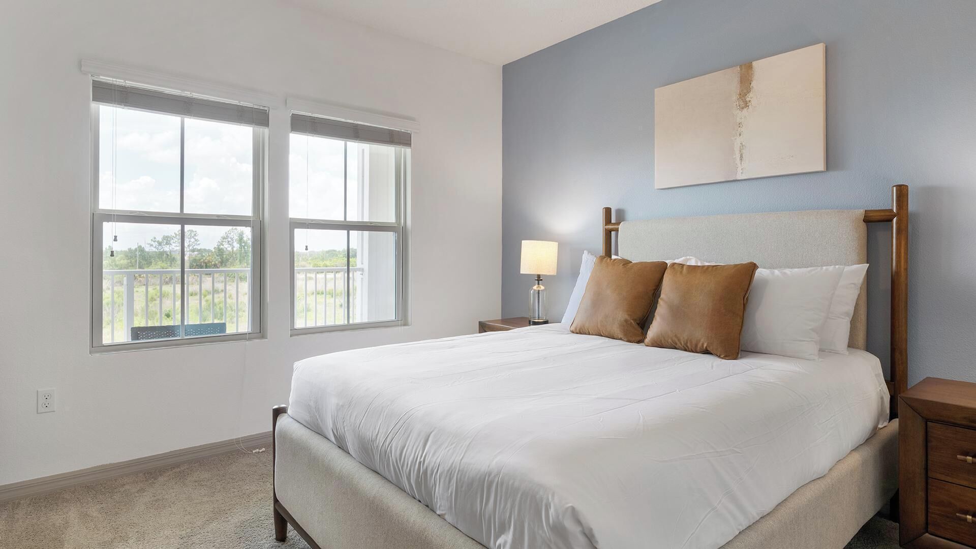 A bedroom with a large bed, two bedside tables, a nightstand, a window with blinds, and a wall-mounted artwork.