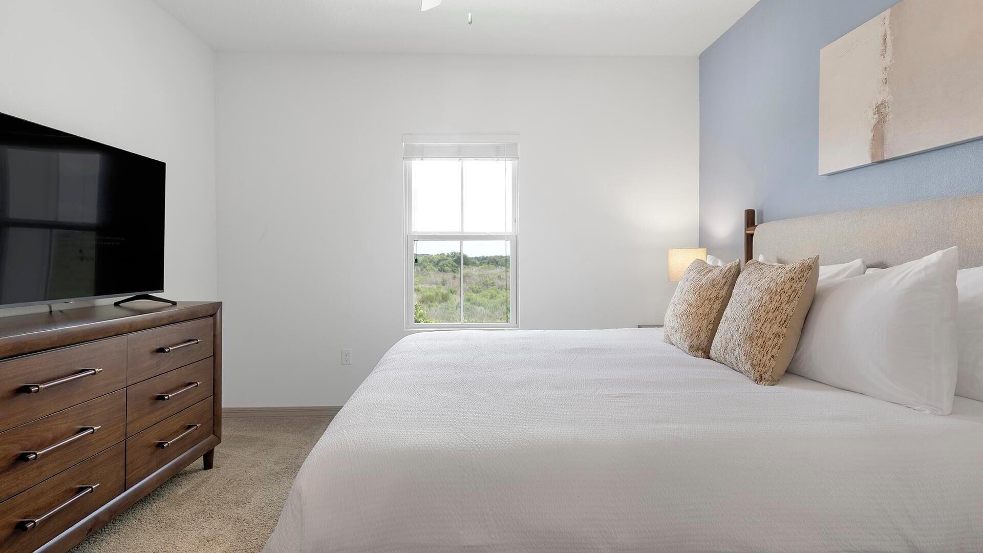 A bedroom with a bed, a dresser, a television, a window, and a painting on the wall.