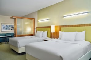 A hotel room with two beds, a desk, a chair, a lamp, and a wall-mounted light fixture.