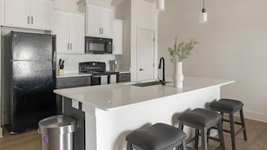 A modern kitchen with white cabinetry, a black refrigerator, a microwave, a stove, and a sink. There are three bar stools and a trash can. - Landing | Chic 1BD, Gym, Clubhouse (Asheville)
