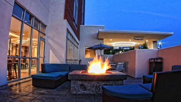 Un área de estar al aire libre con un hogar de piedra, asientos acolchados y un muro de vidrio que da a un comedor interior.