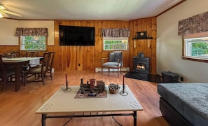 Una habitación acogedora con paredes de madera, un televisor instalado en la pared, chimenea, una mesa de comedor con sillas y un sofá.