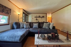 A living room with a sectional sofa, a small table with a lamp, a decorative train, and a wall-mounted shelf with a clock and a bicycle.