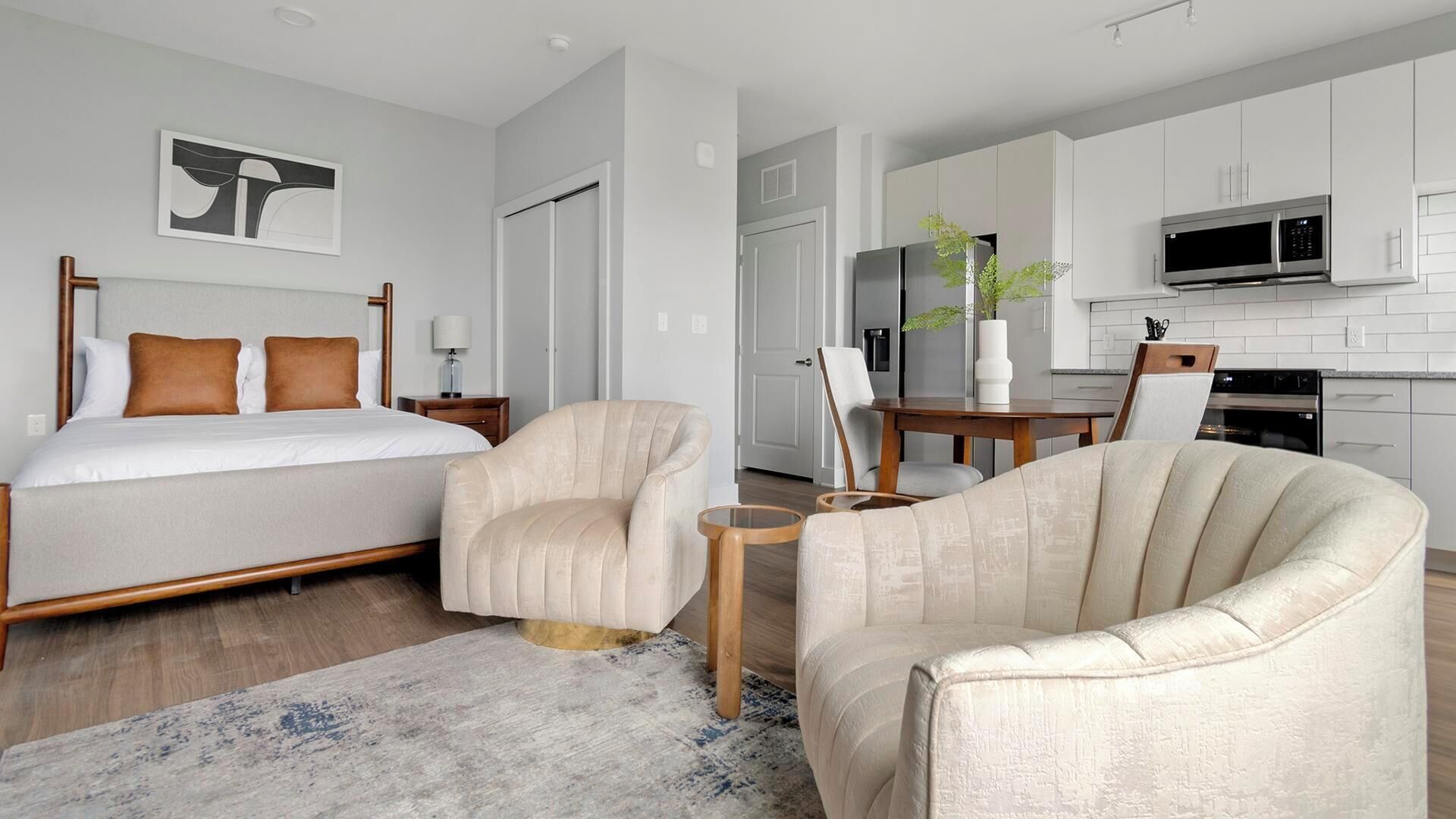 Una habitación de hotel moderna con una cama, dos sillones, una mesita, un escritorio, una silla, una mesa de comedor, un microondas, un refrigerador y una planta en maceta.