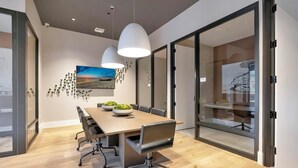 A modern meeting room with a wooden table, black chairs, pendant lights, and a cityscape painting on the wall.