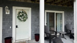 A porch with a white door, a wreath, two Adirondack chairs, and a red bucket.