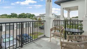 A balcony with a view of parked cars, a wicker chair, a table, and a building with a tiled roof. - Landing | Incredible 1BD, Gym (Sun City Center)