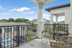 A balcony with a view of parked cars, a wicker chair, a table, and a building with a tiled roof. - Landing | Spectacular 1BD, Gym (Sun City Center)