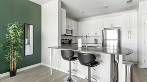 A modern kitchen with white cabinets, stainless steel appliances, and a bar area with two stools. - Landing | Incredible 1BD, Gym (Sun City Center)