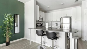 A modern kitchen with white cabinets, stainless steel appliances, and a bar area with two stools. - Landing | Spectacular 1BD, Gym (Sun City Center)