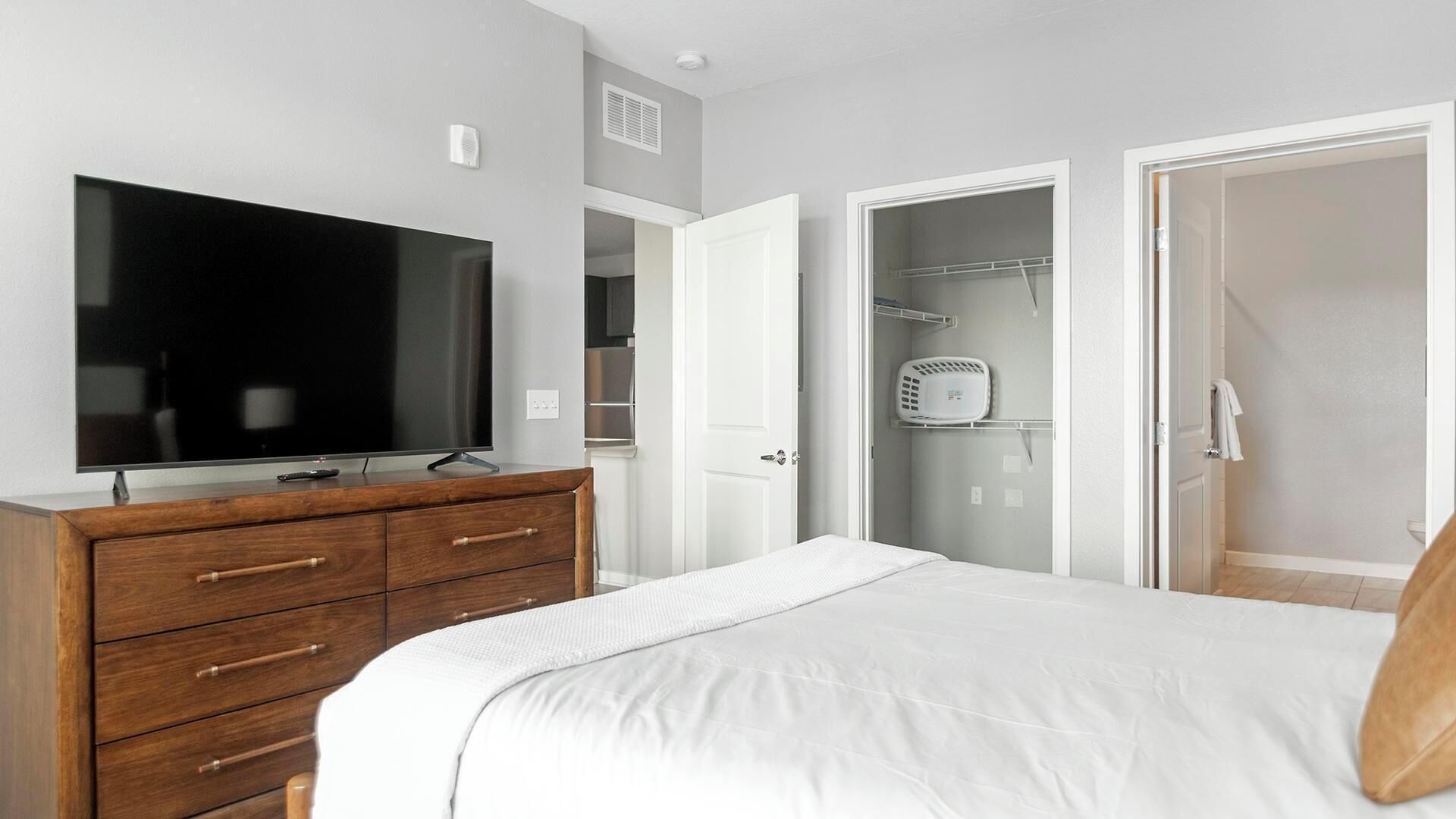 A bedroom with a TV, wooden dresser, white bed, open closet, and a shower area.
