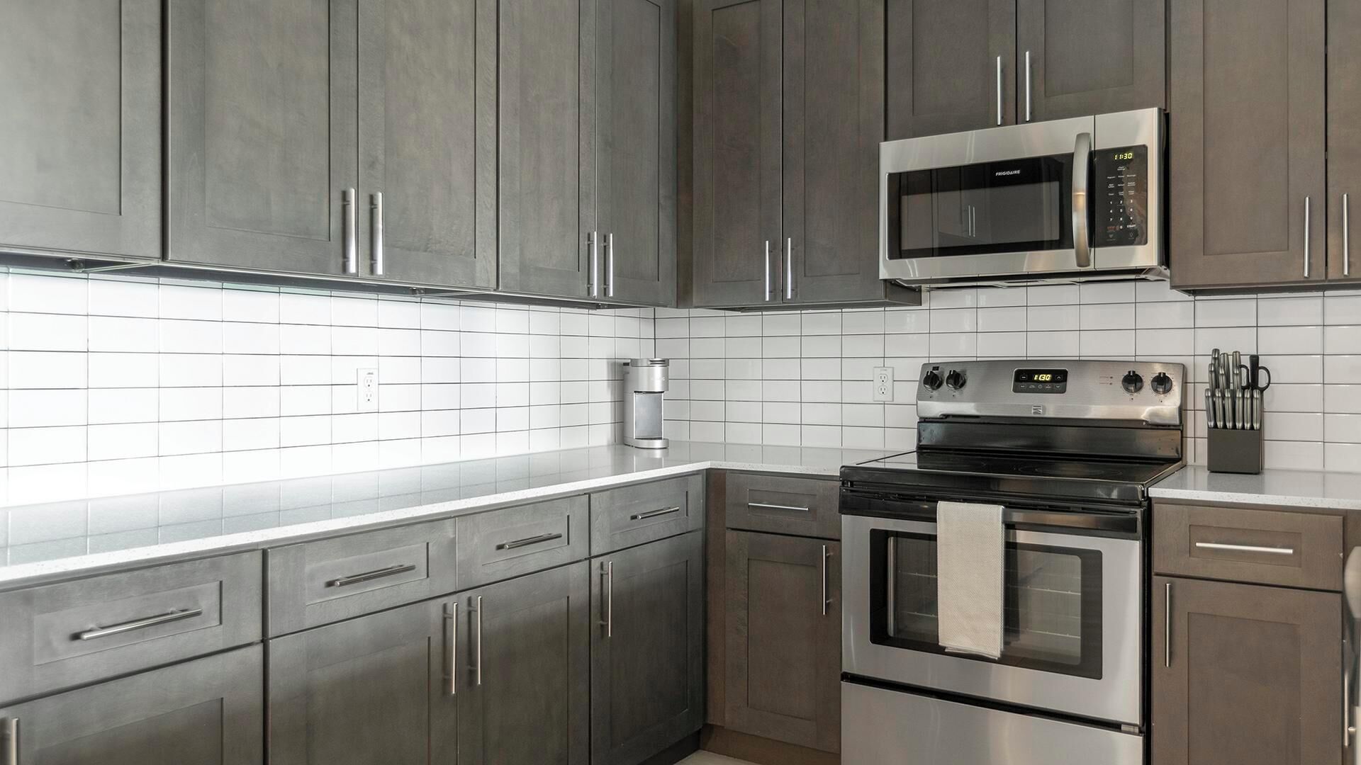 A kitchen with stainless steel appliances, grey cabinets, and white tiled backsplash.