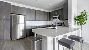Cozinha moderna com armários cinza, eletrodomésticos em aço inoxidável, uma ilha central com banquetas e uma planta em vaso.