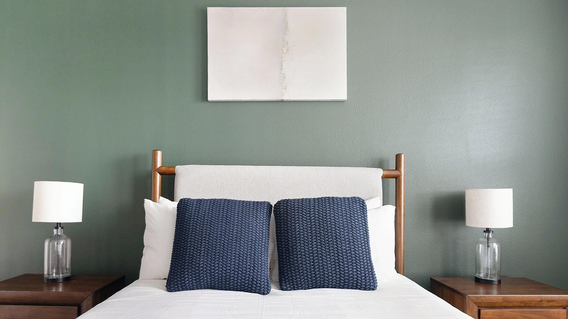 A bed with a wooden headboard, two bedside tables with lamps, and a plain wall with a framed picture.