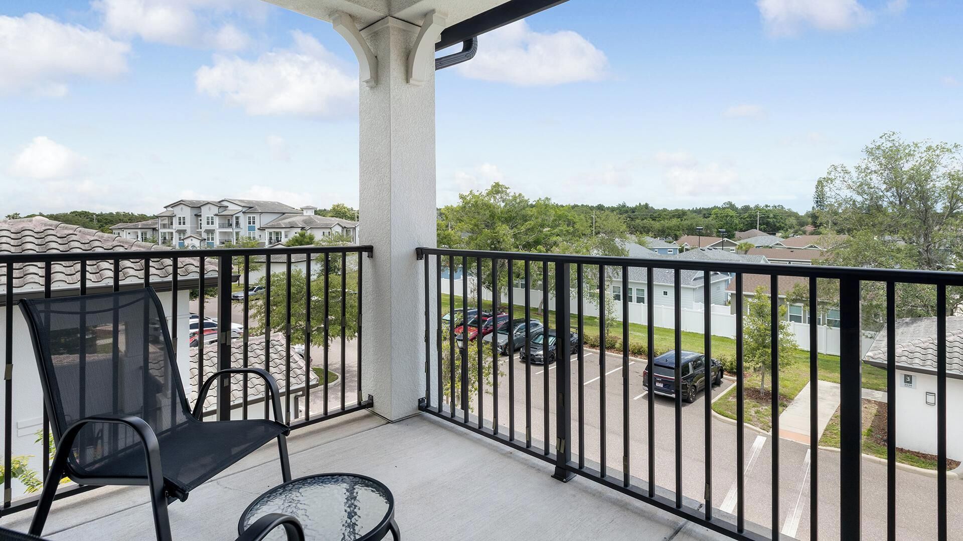A balcony with a chair and table, a view of a parking lot, and residential buildings in the background.