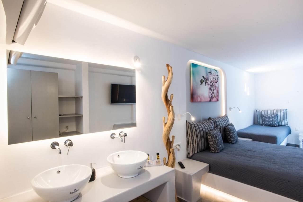 A modern hotel room with a large mirror, two sinks, a bed with patterned bedding, a mounted TV, and a decorative wooden piece.