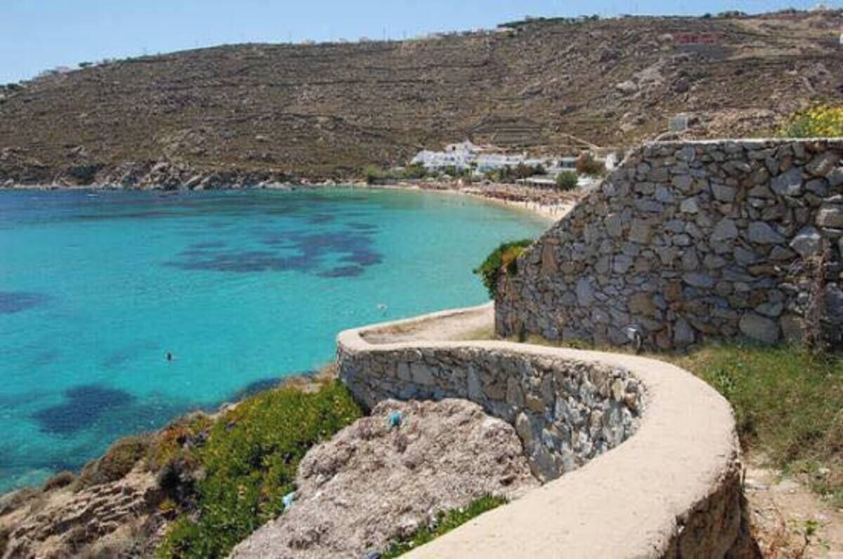 A coastal landscape with a stone wall, clear turquoise water, and a distant building on a hill.