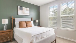 A bedroom with a bed, two bedside tables, a nightstand, a window with blinds, and a wall-mounted artwork.