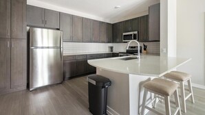 A modern kitchen with dark cabinets, stainless steel appliances, a large island with stools, and a trash bin. - Landing | Spectacular 2BD, Gym (Sun City Center)
